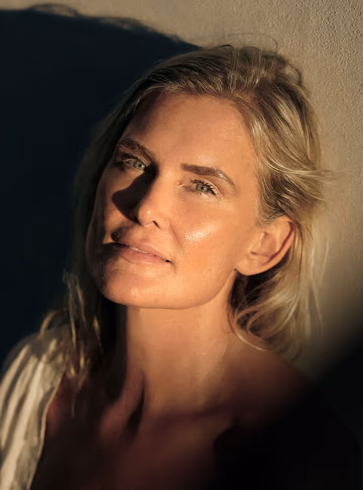 Close-up portrait of a woman with blonde hair and green eyes, softly lit with warm sunlight casting shadows on her face and background.