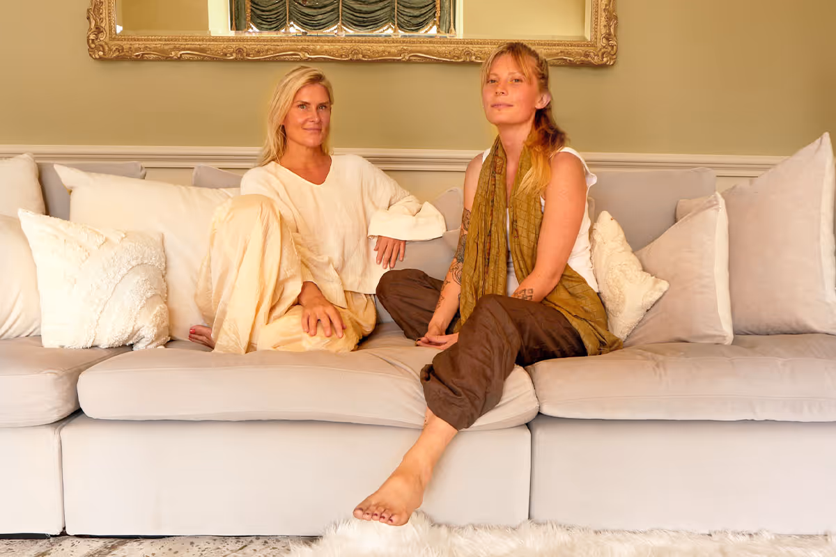 Two women sitting barefoot on a beige sofa in a living room with decorative pillows and a large ornate mirror on the wall behind them.