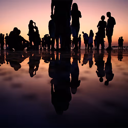 Silhouettes of people standing and walking with their reflections on wet ground during a sunset.