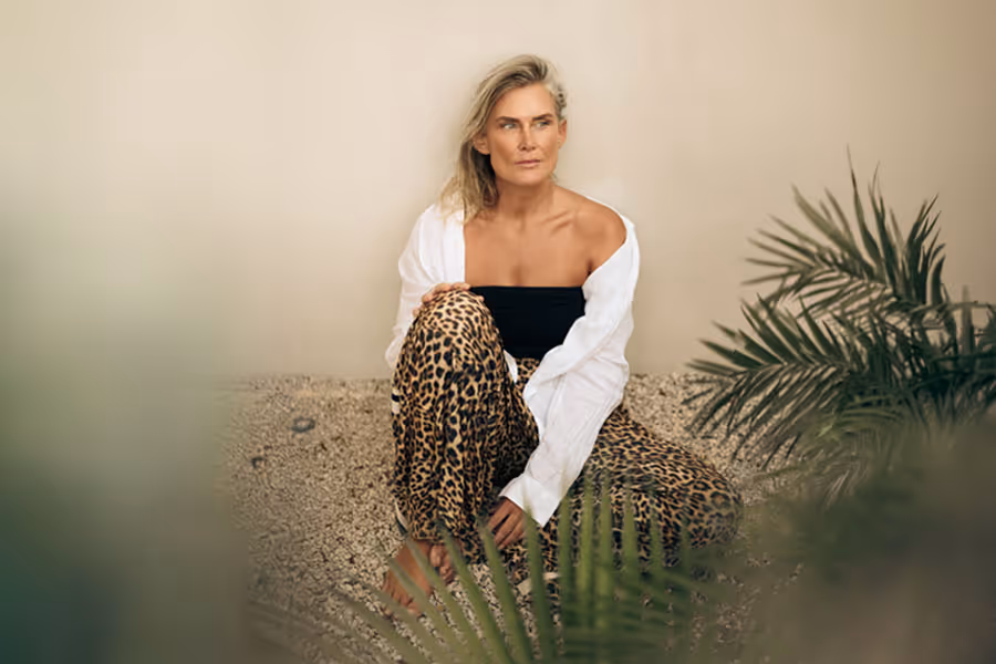Woman sitting barefoot on gravel floor between green plants, wearing leopard-print pants, black top, and white open shirt.