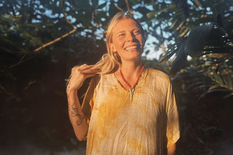 Smiling woman with blonde hair wearing a yellowish-beige shirt, red necklace, and tattoo on her forearm, standing outdoors with greenery in the background.