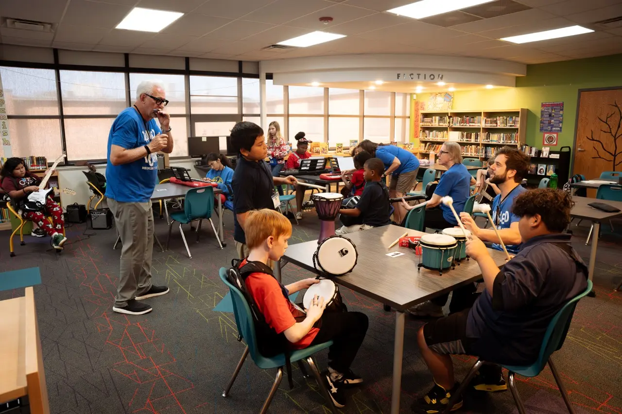 Children and adults playing drums and guitar in a brightly lit library room.