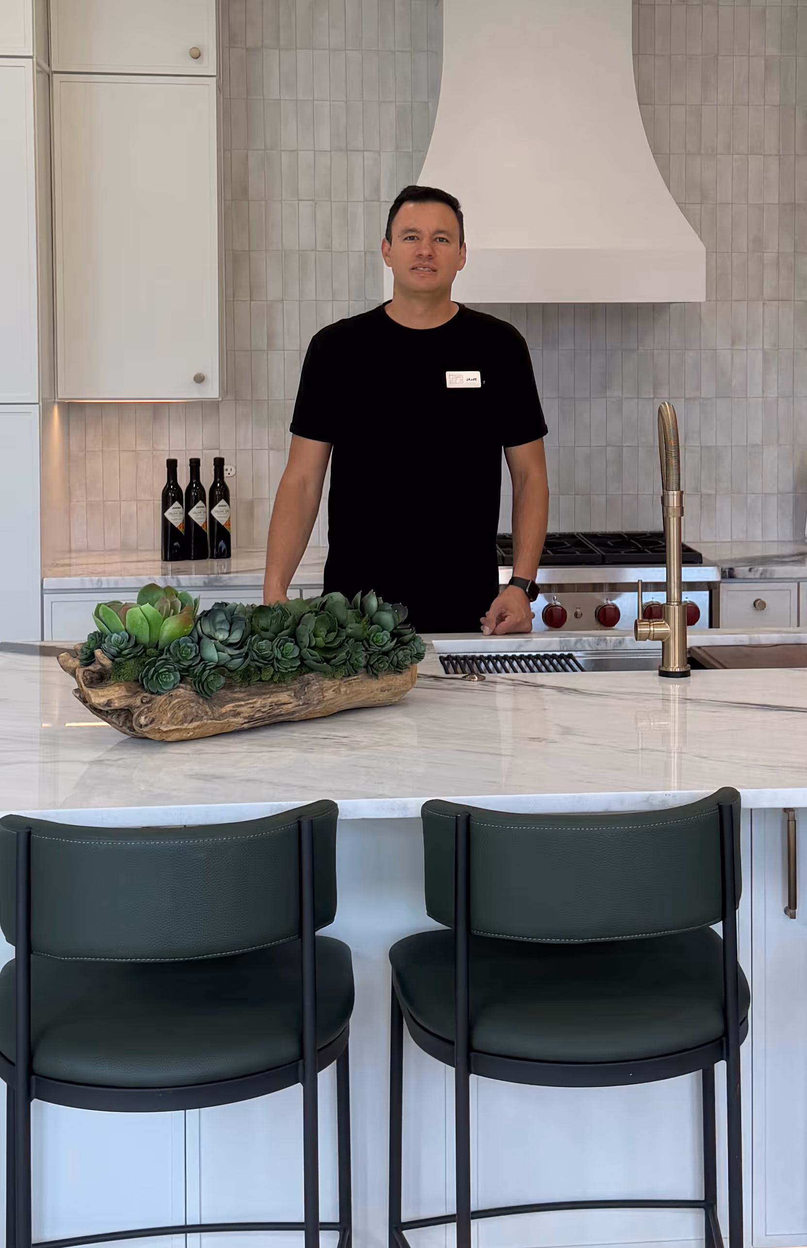 Man in black shirt stands behind kitchen island with succulent centerpiece and two green bar stools.