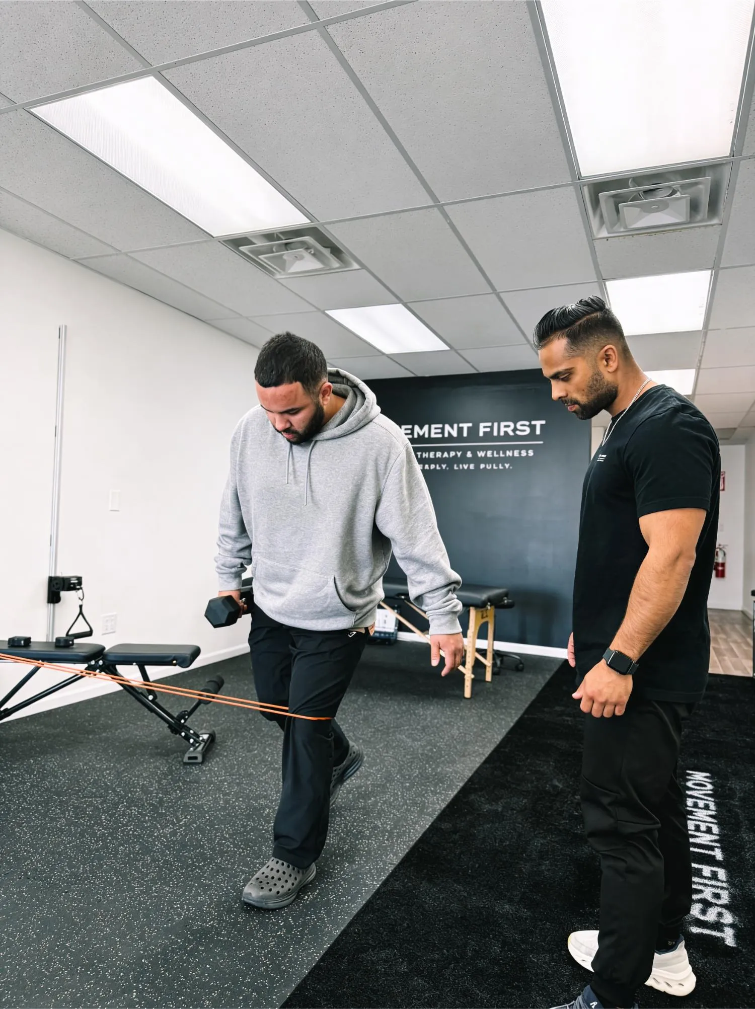 Man in gray hoodie exercises with resistance band and dumbbell under trainer's supervision in gym.