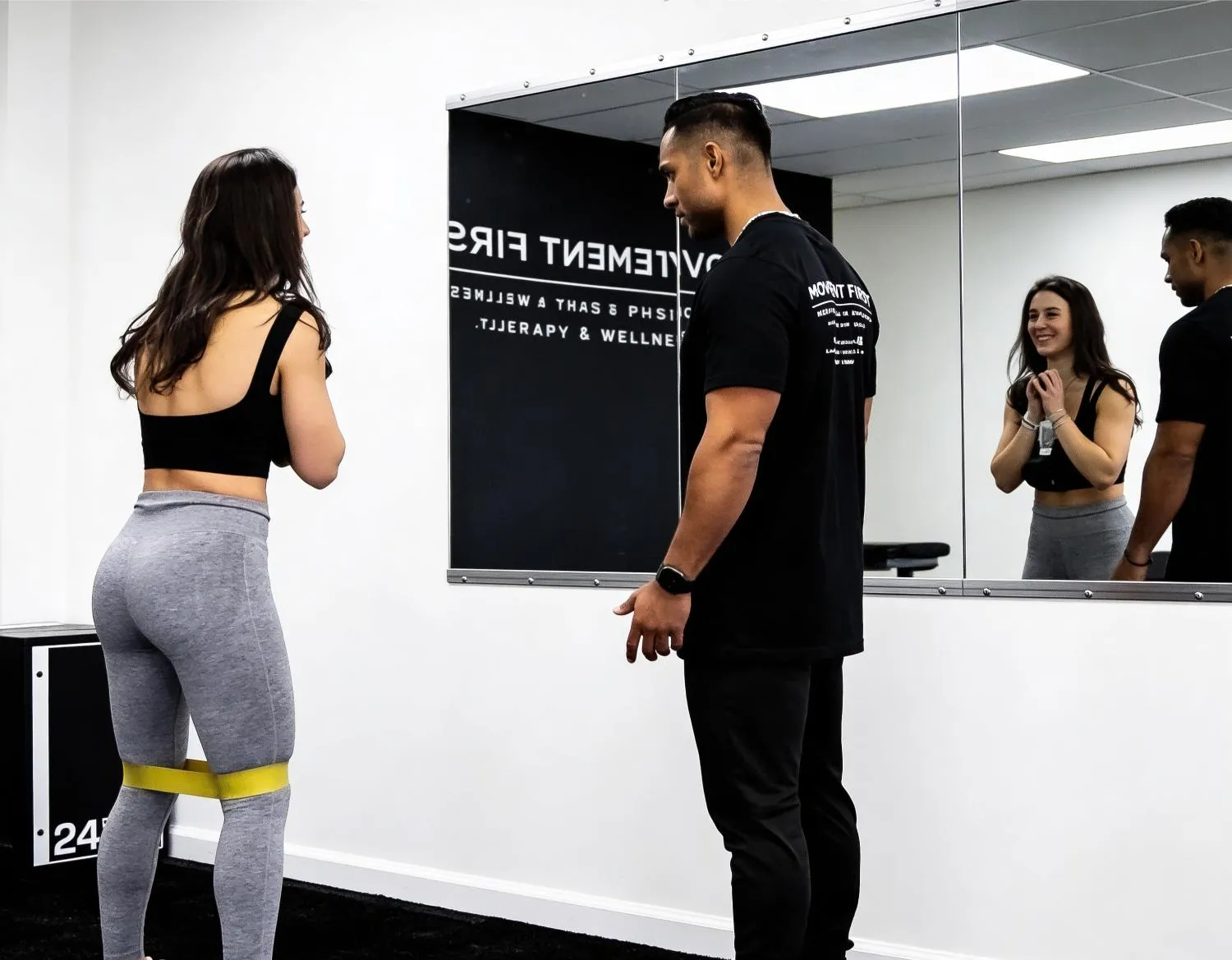 Woman exercises with yellow resistance band on legs while trainer watches in gym with mirror.