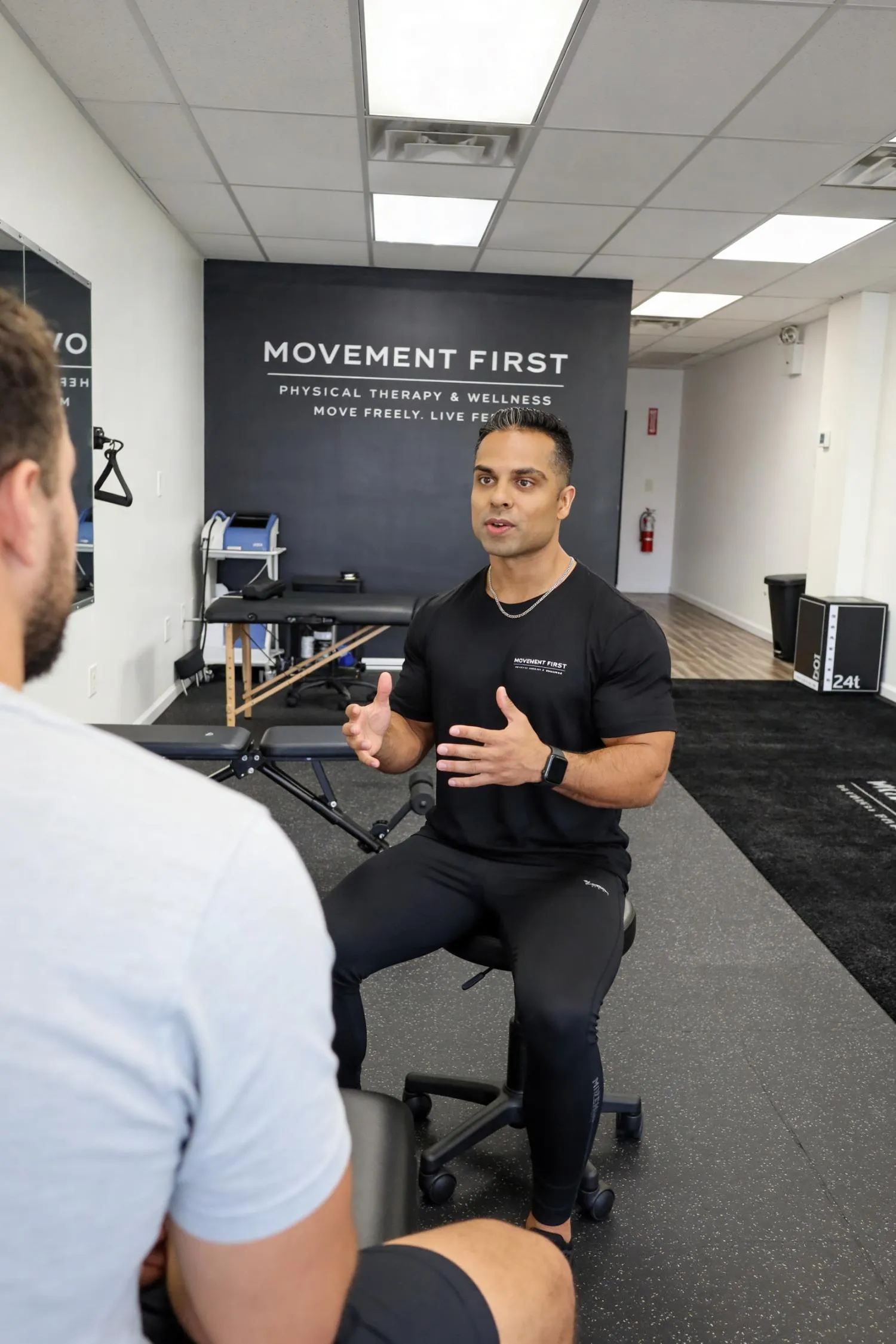 Physical therapist in black speaks with a seated patient in a fitness and wellness clinic.