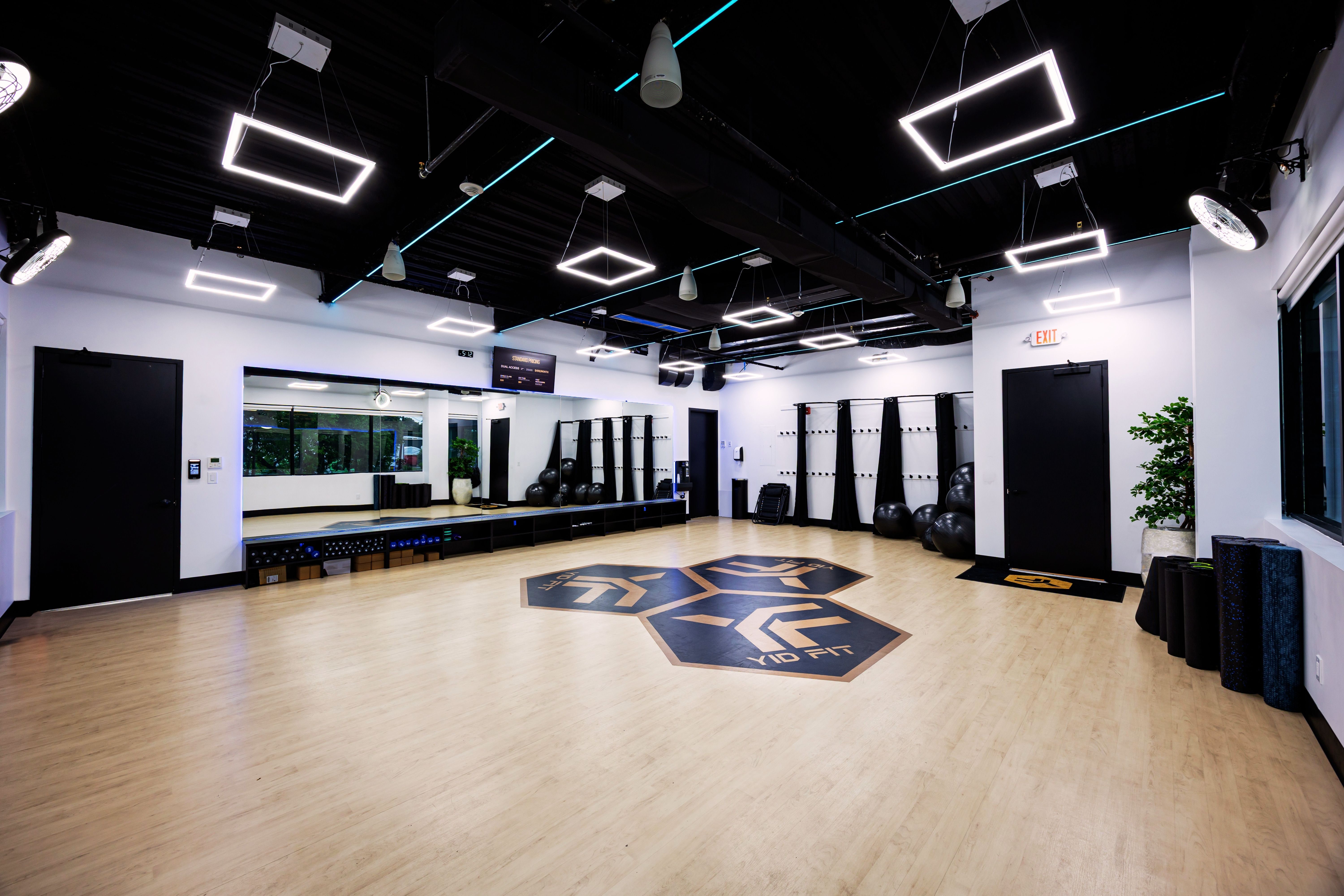 Modern gym room with stationary bikes, treadmills, elliptical machines, weights, large windows, and distinctive hexagonal ceiling lights.