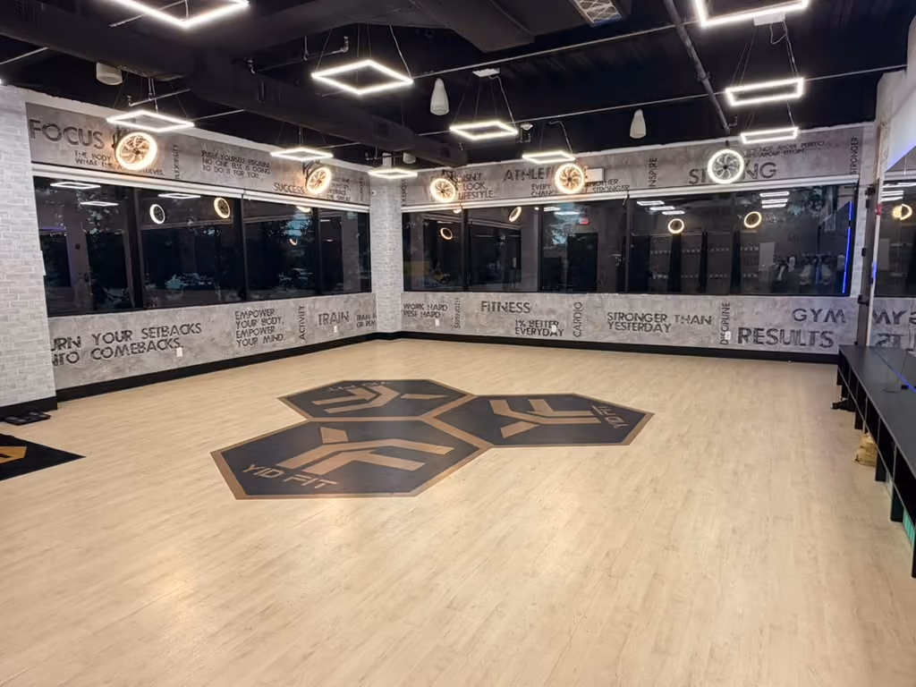 Modern gym room with stationary bikes, treadmills, elliptical machines, weights, large windows, and distinctive hexagonal ceiling lights.