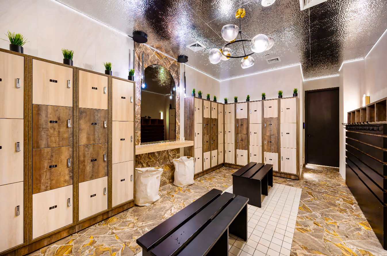 Modern locker room with light wood lockers, black benches, marble floor, and plants on top of lockers.