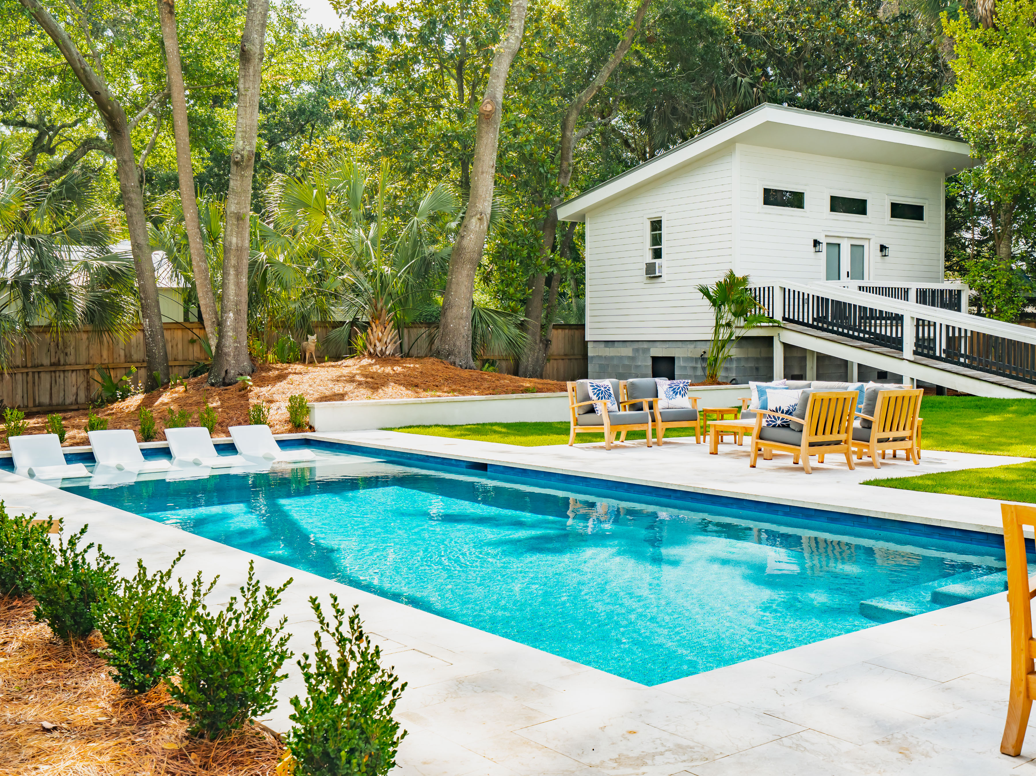 A rectangular swimming pool with submerged lounge chairs, a stone patio, and a white guest house in the background.