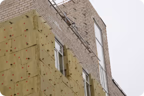 Gebäudewand mit Dämmplatten vor der Fertigstellung und zwei Fenstern, auf der anderen Seite Sicht auf Ziegelmauerwerk.