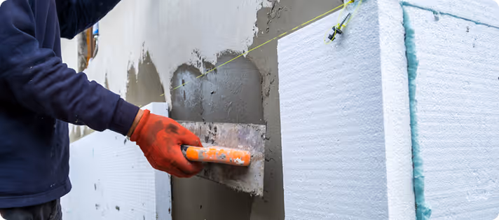 Arbeiter trägt rote Handschuhe und glättet Zement mit einem Glätter an einer Hauswand mit Dämmplatten.