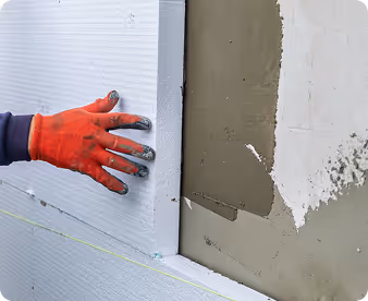 Hand in orange glove pressing insulating foam board onto wall with fresh cement plaster.