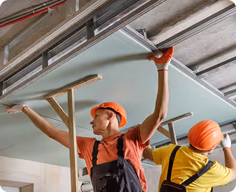 Zwei Bauarbeiter mit orangenen Helmen montieren eine Deckenplatte in einem Rohbau.