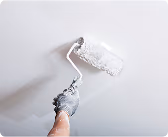 Hand mit Handschuh, die mit einer Farbrolle eine weiße Decke streicht.