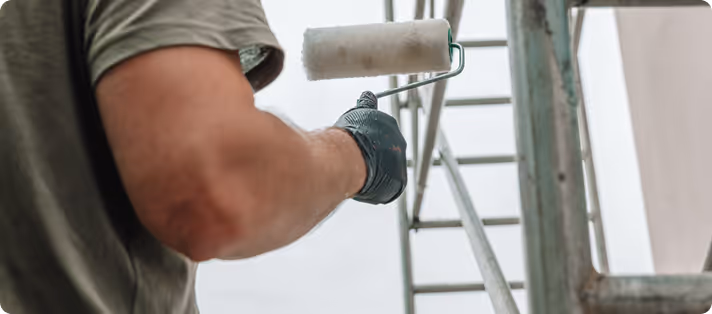 Person mit schwarzem Handschuh hält eine weiße Farbrolle vor einem Metallgerüst und einer Leiter.