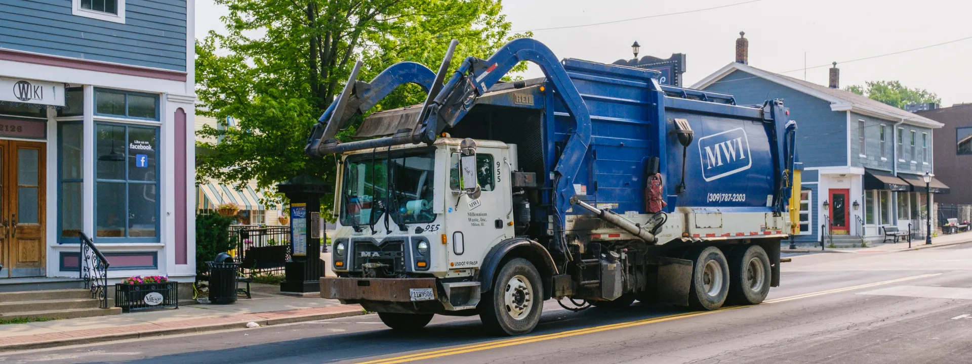 Blue and white front-load garbage truck driving on a street in a small town with buildings and trees.