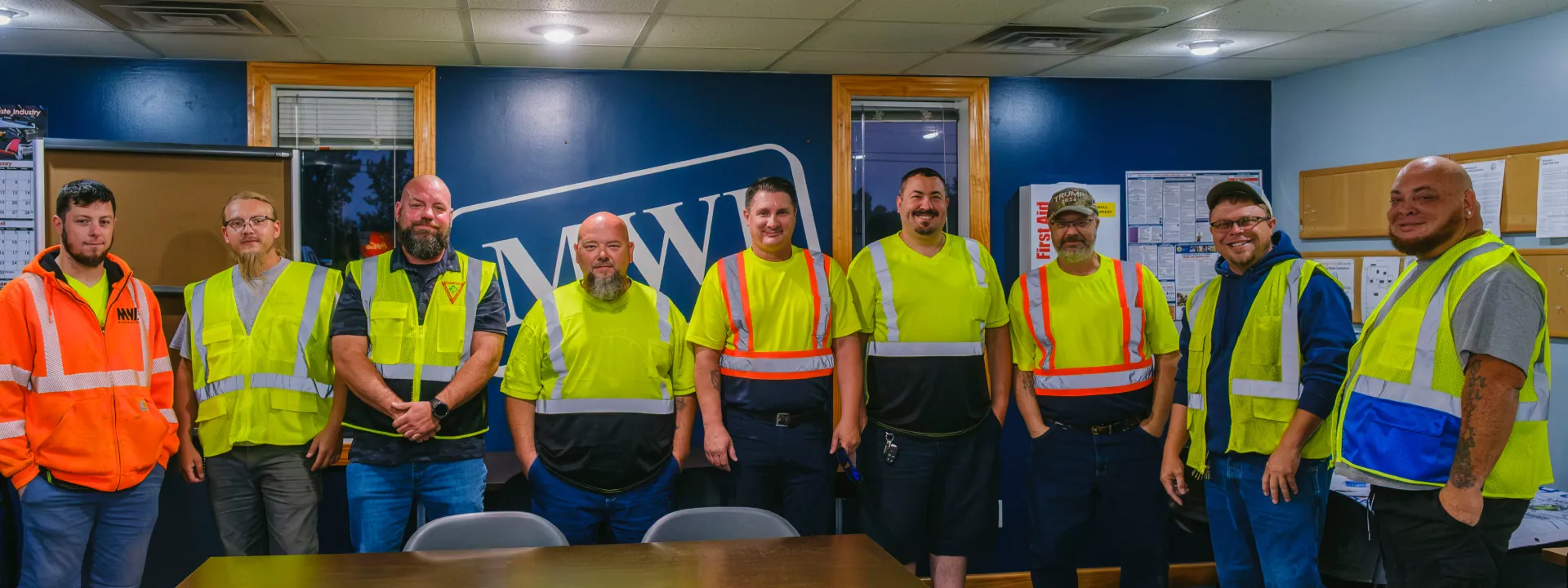 Group of nine waste service workers in safety vests standing indoors against a blue wall with MW logo.