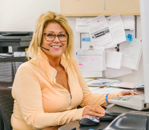 Woman with headset sitting at office desk smiling while using a computer mouse and keyboard.