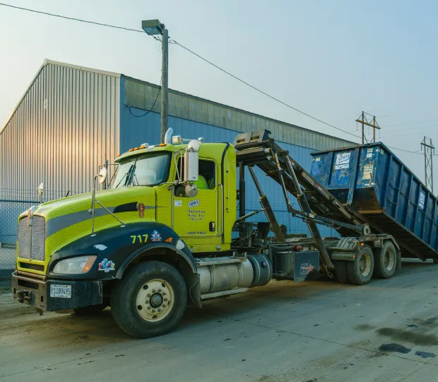 Green and black roll-off truck transporting a large blue dumpster for construction waste removal.