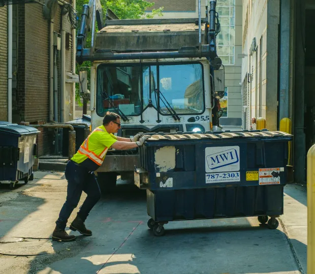 Worker in safety vest pushing a large dumpster behind a truck in an alley between buildings.