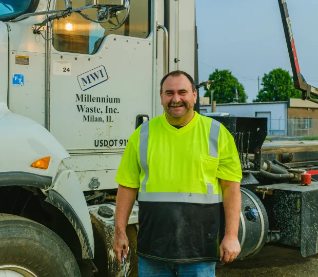 Smiling sanitation worker in a yellow safety shirt standing in front of a Millennium Waste truck.