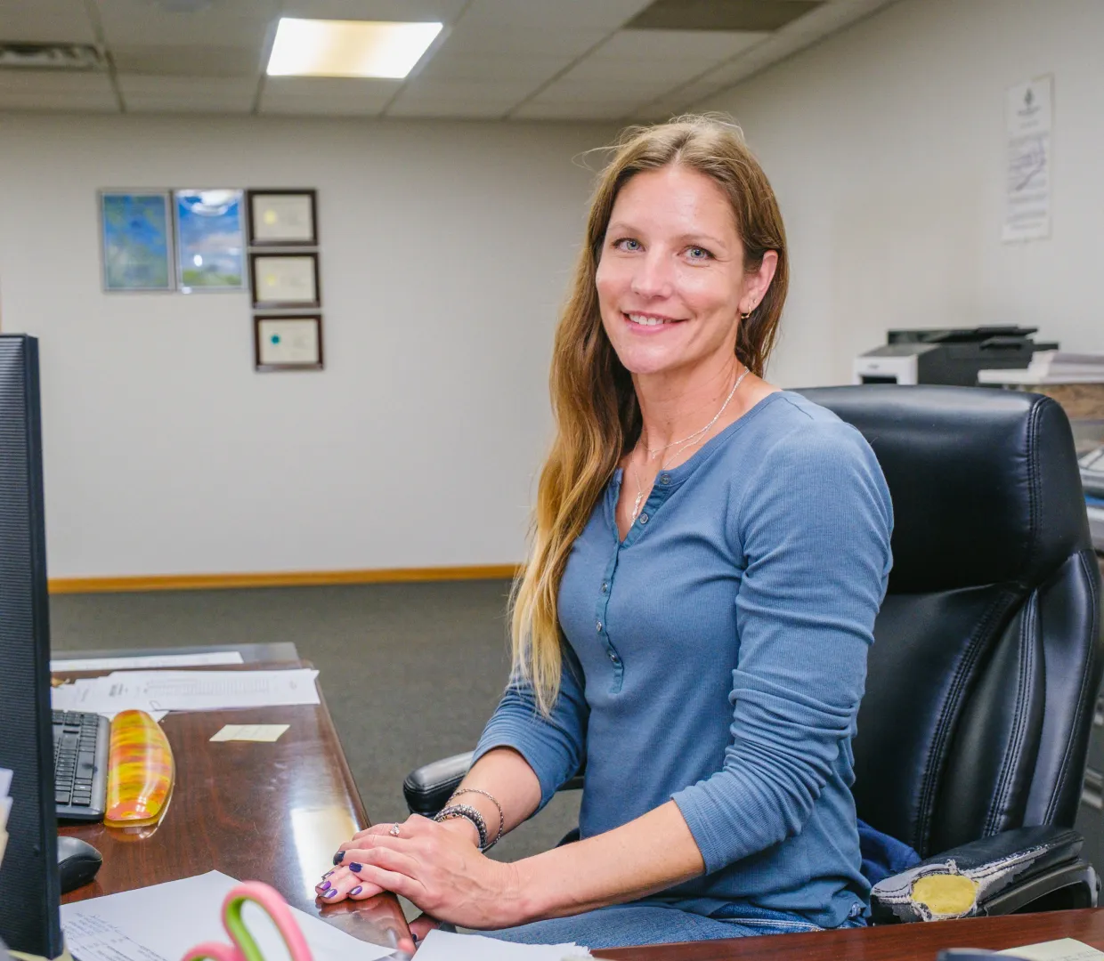 Smiling woman with long hair seated at an office desk with computer and papers.