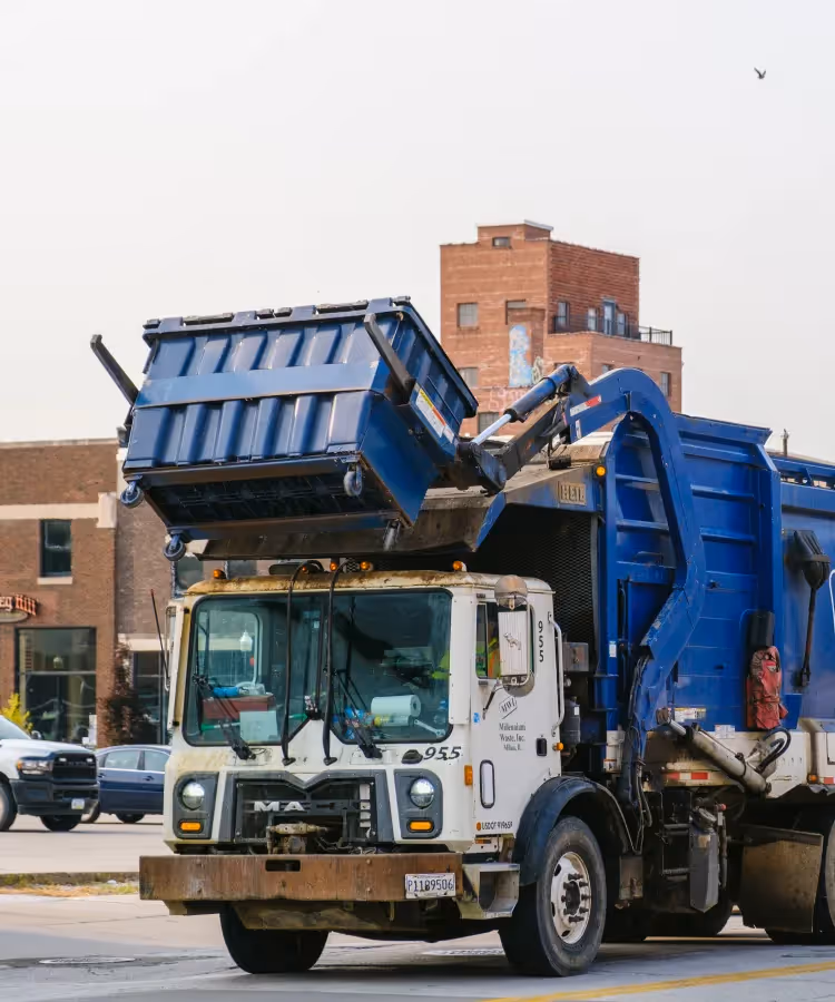 White and blue garbage truck lifting a blue dumpster on a city street with buildings in background.