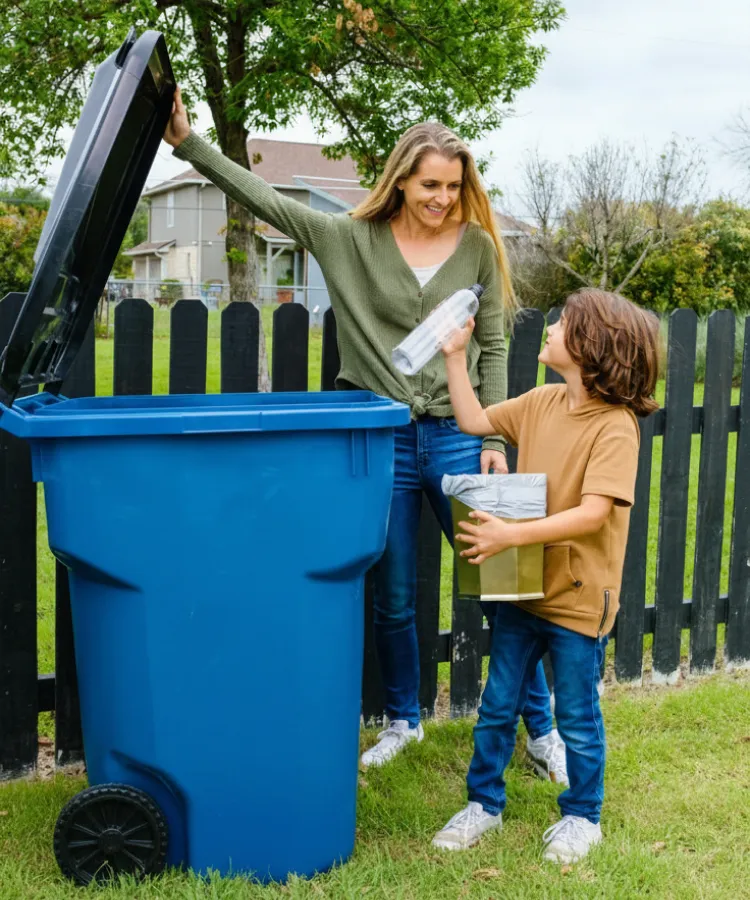 Woman helps boy recycle a plastic bottle into a blue recycling bin outdoors by a black fence.