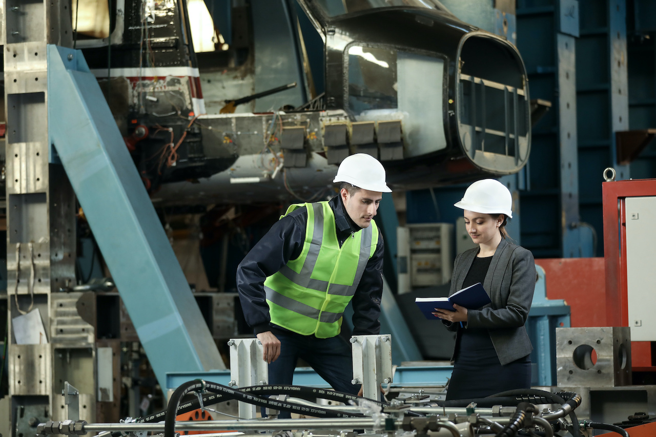 Two engineers wearing white hard hats inspecting machinery in an industrial setting, one reading from a notebook and the other leaning on equipment.