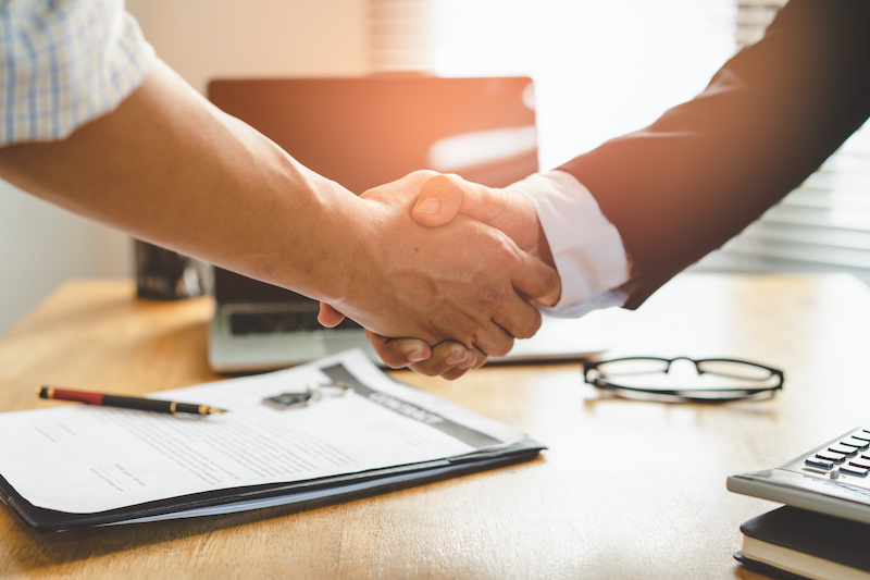 Two people shaking hands over a desk with documents, glasses, and a calculator, symbolizing agreement or partnership.