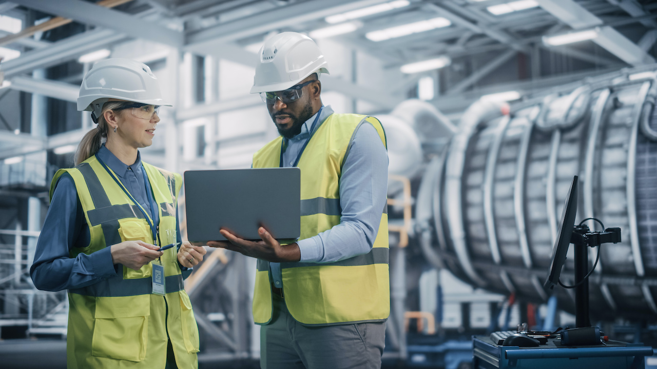 Two engineers wearing helmets and reflective vests discussing work while looking at a laptop in an industrial facility with large machinery.