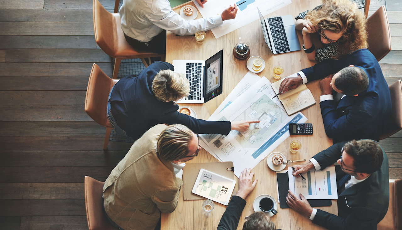 Six professionals collaborating around a wooden table with laptops, documents, and drinks, discussing plans and data charts.