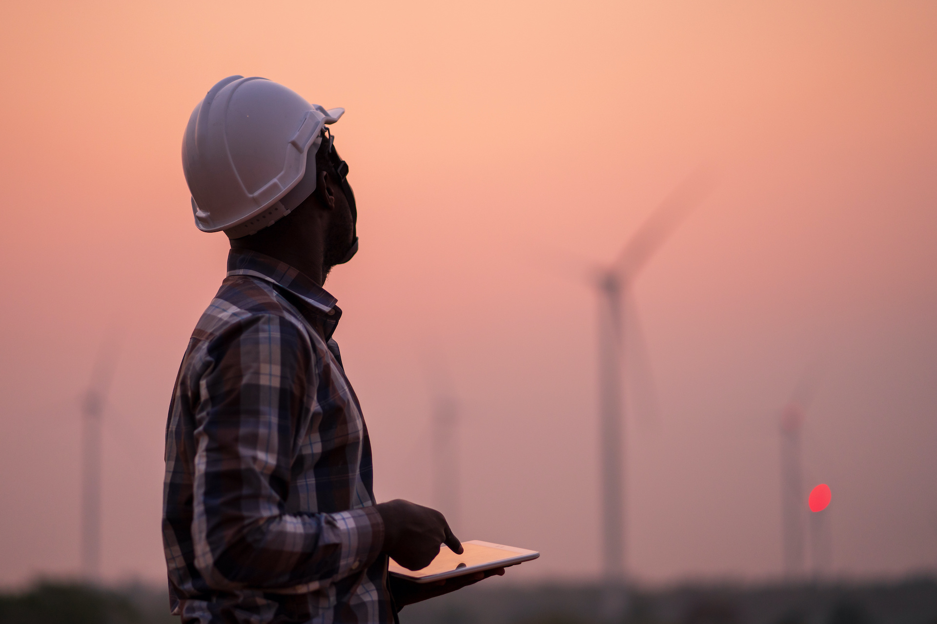 Engineer wearing a white hard hat and plaid shirt using a tablet while inspecting wind turbines at sunset.