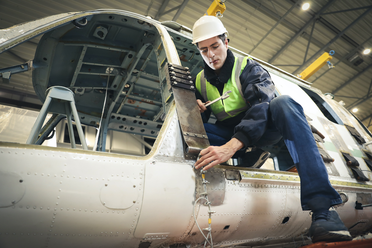 Technician in a hard hat and safety vest repairing the exterior of a disassembled helicopter inside a hangar.