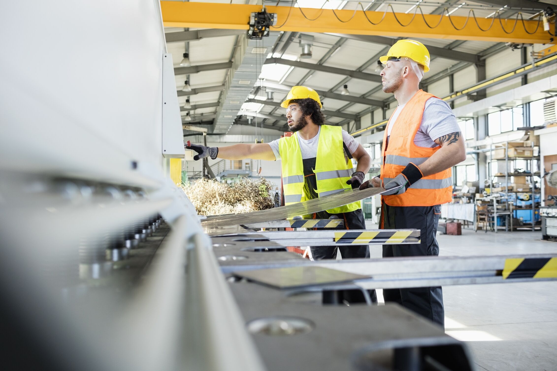 Two workers in safety vests and helmets operating metalworking machinery in a factory.