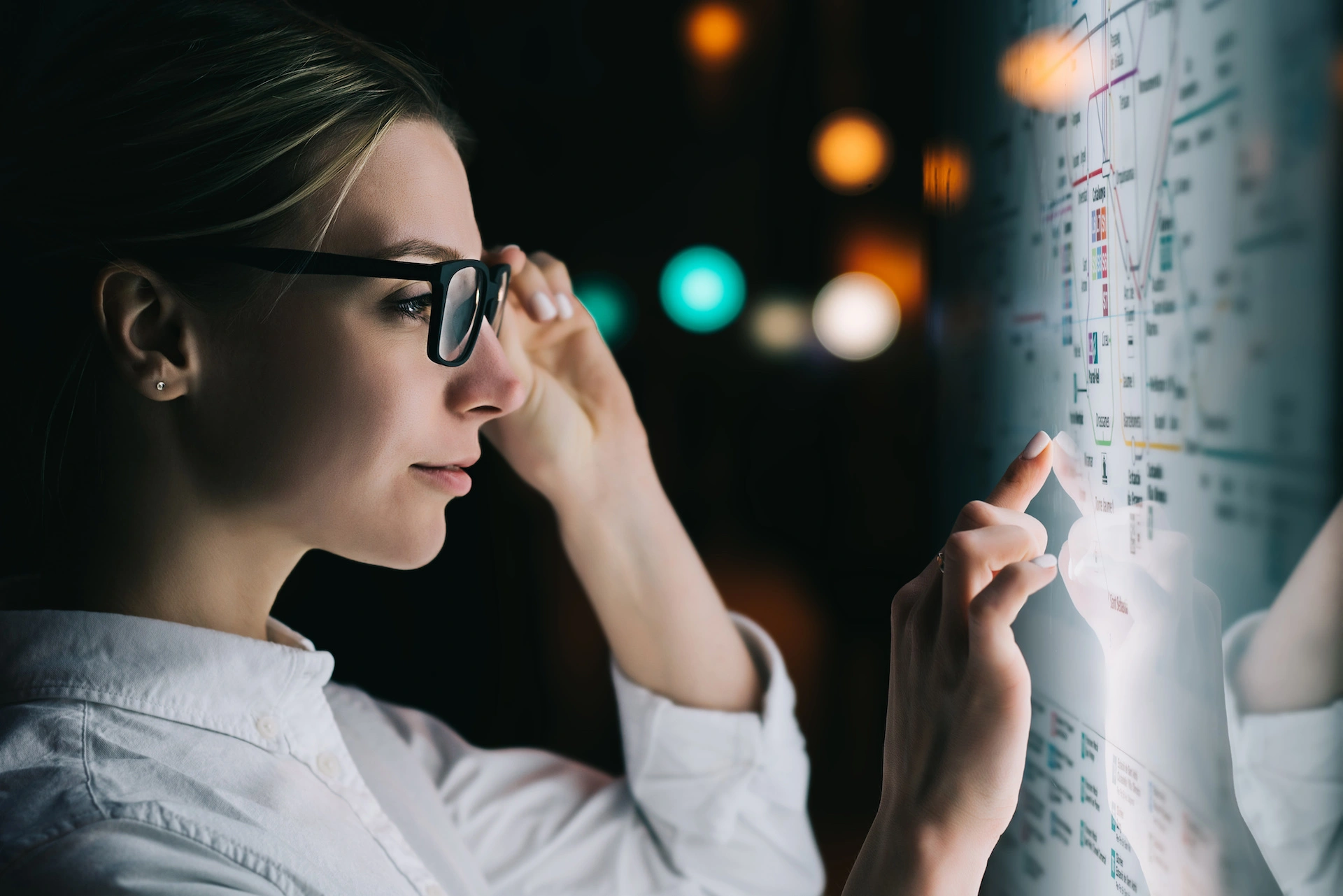 Woman in glasses interacting with a large digital touch screen displaying a map with illuminated points and lines.