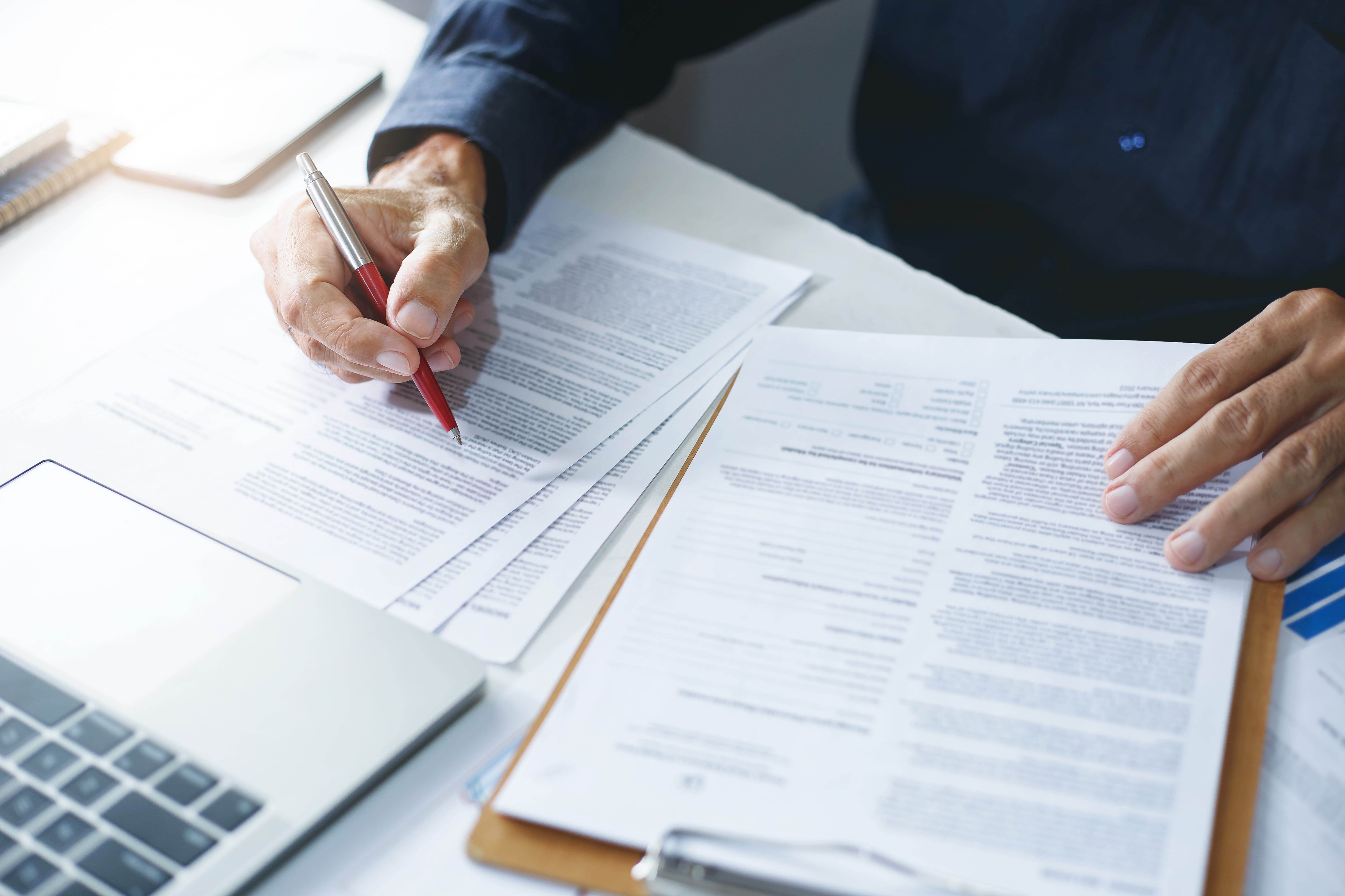 Hands of a person reviewing and marking documents with a red pen beside a laptop on a white desk.
