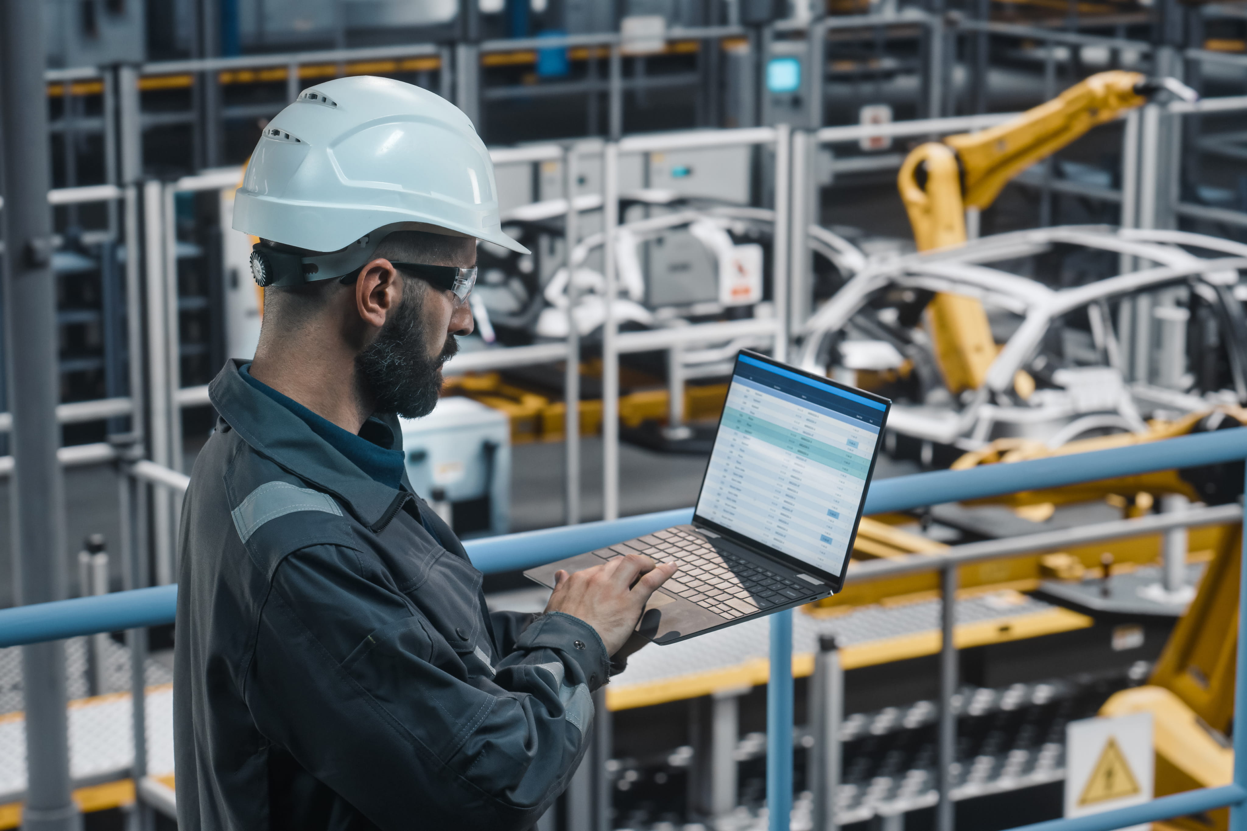 Industrial worker wearing a hard hat and safety glasses using a laptop in a factory with robotic machinery and car frames.