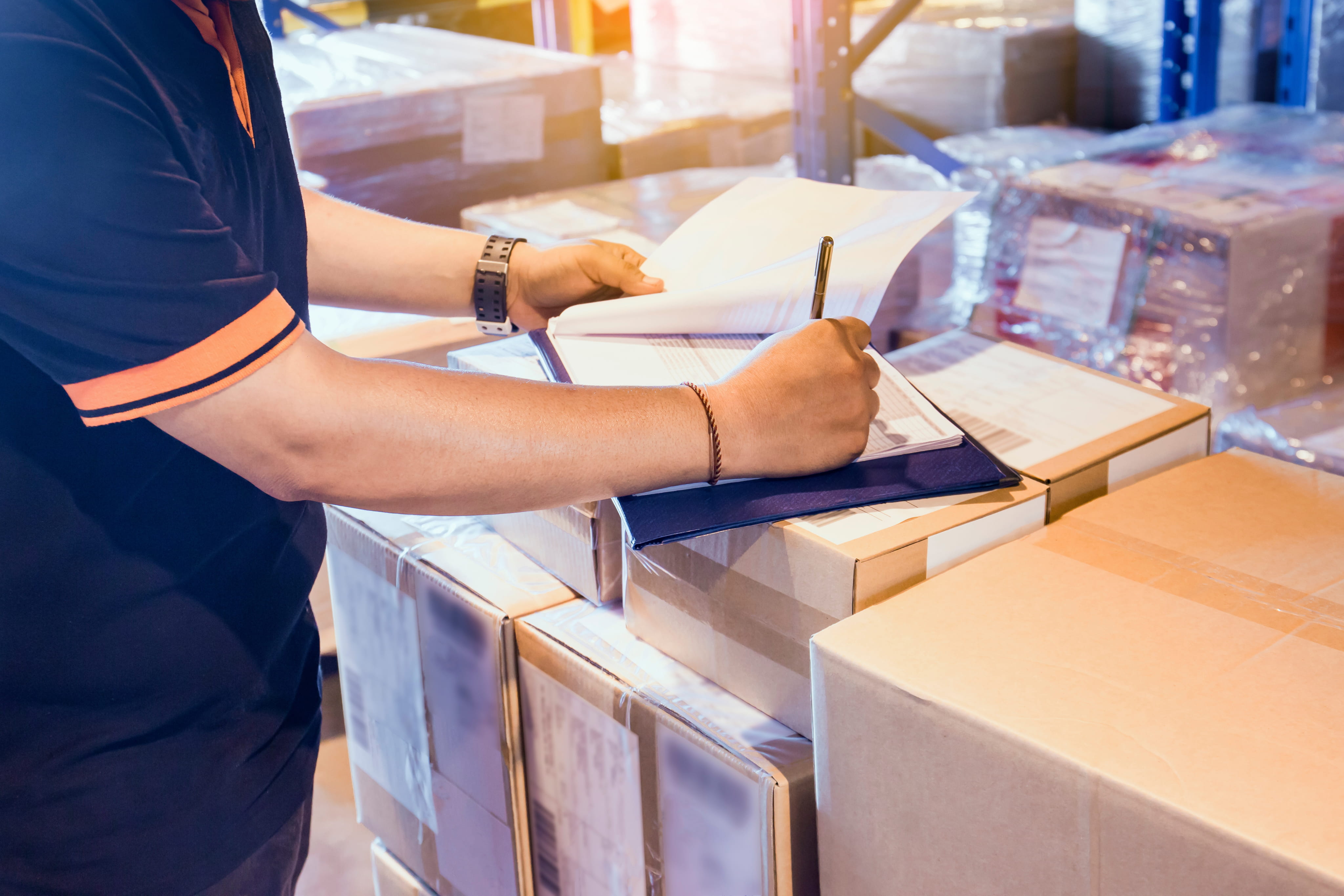 Person in black shirt checking and signing documents on a clipboard over stacked cardboard boxes in a warehouse.