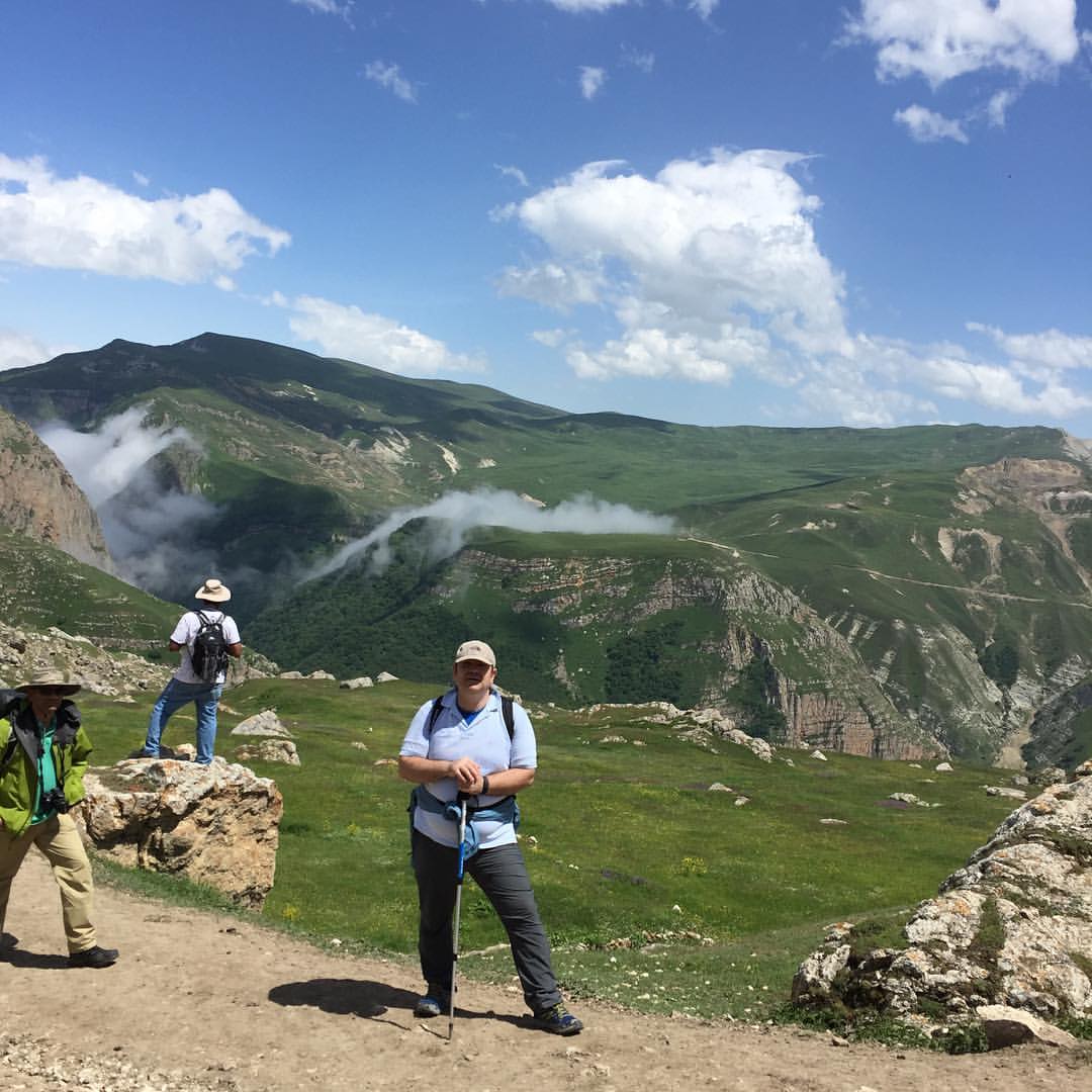 A hiker pauses on the trail above the high Caucasus valley near Qırız, two companions looking out over green ridges and drifting cloud — from the founder's archive, 2016.