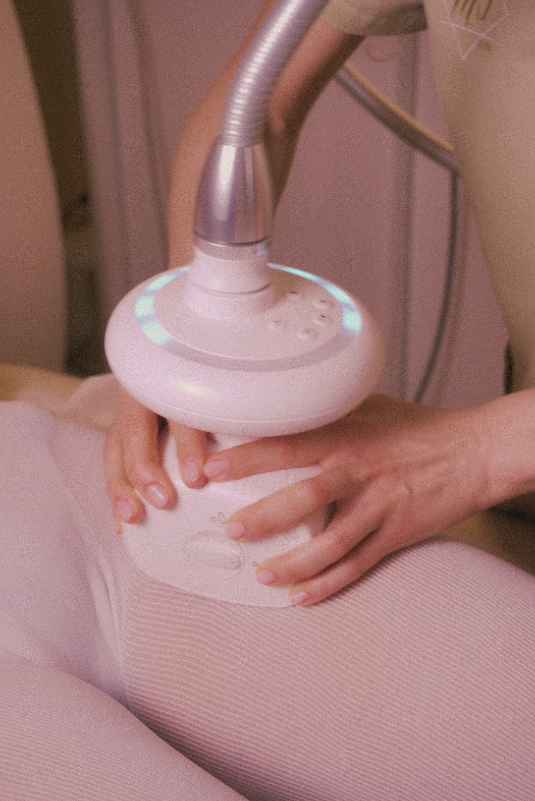 Close-up of hands holding a white body contouring machine on a person's thigh dressed in light pink ribbed fabric.