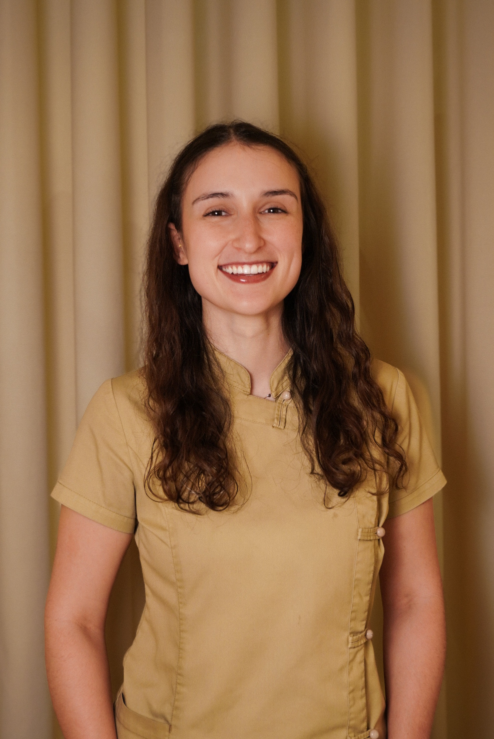 Smiling woman with long curly hair wearing a beige short-sleeve uniform in front of beige curtains.
