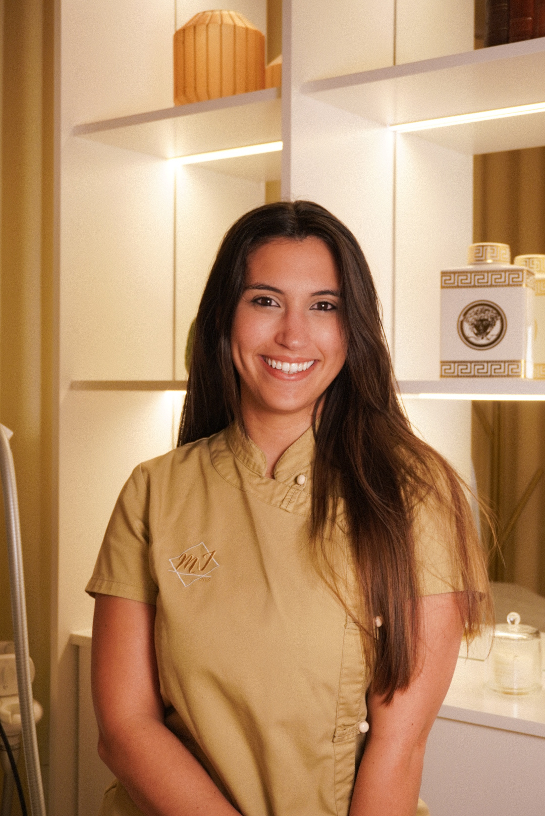 Smiling woman with long dark hair wearing a beige uniform standing in a warmly lit room with shelves in the background.