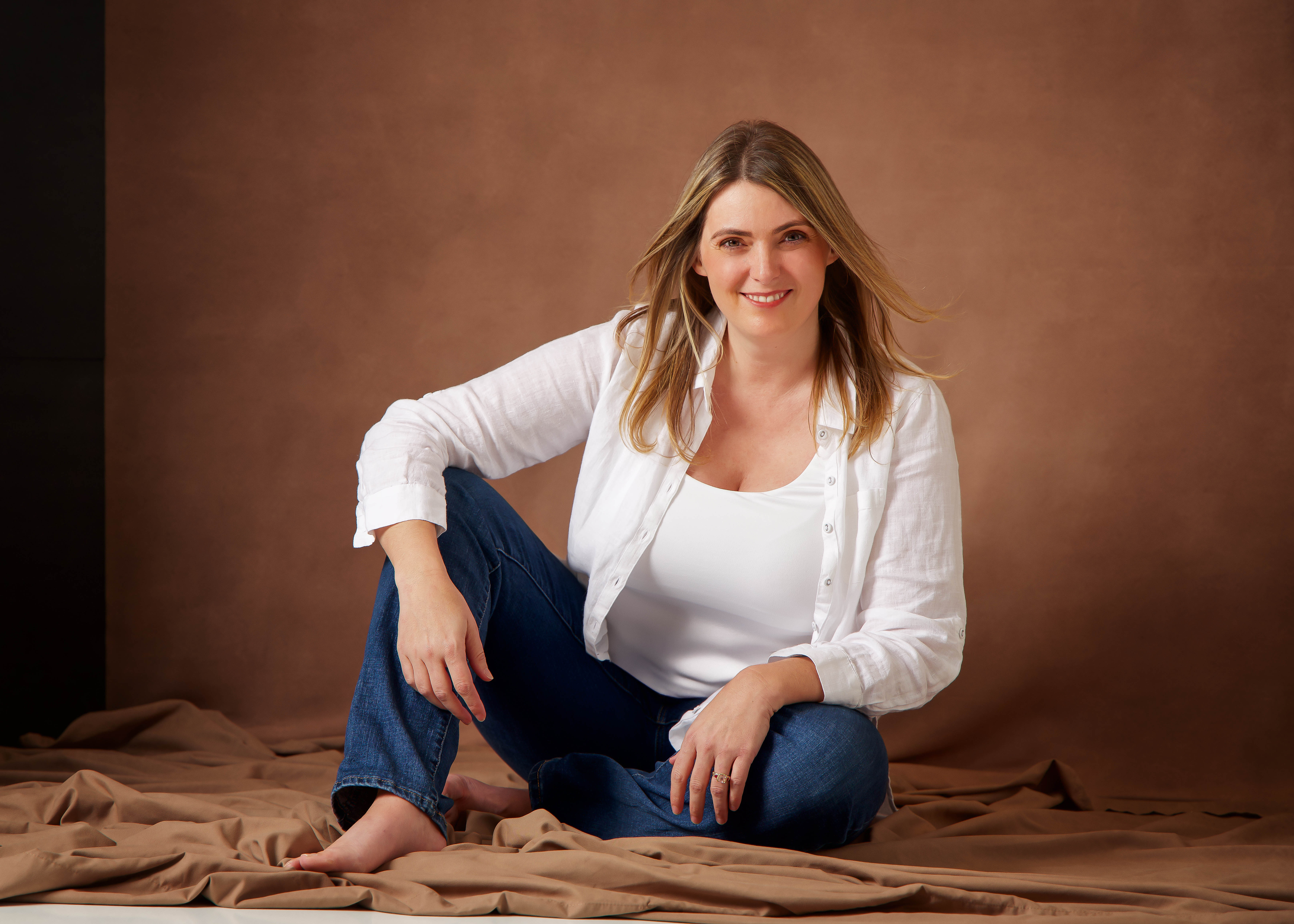 Smiling woman with blonde hair wearing white shirt and blue jeans sitting barefoot on brown fabric.