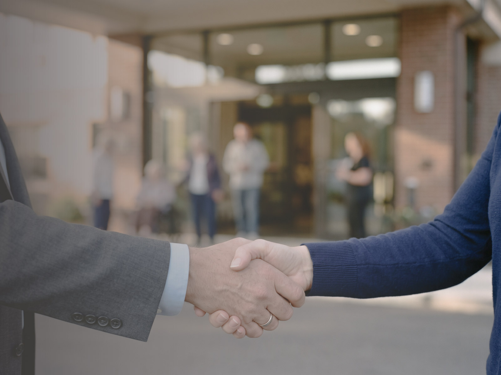 Owner and buyer shake hands to sell an assisted living facility with a glass-door entrance behind them