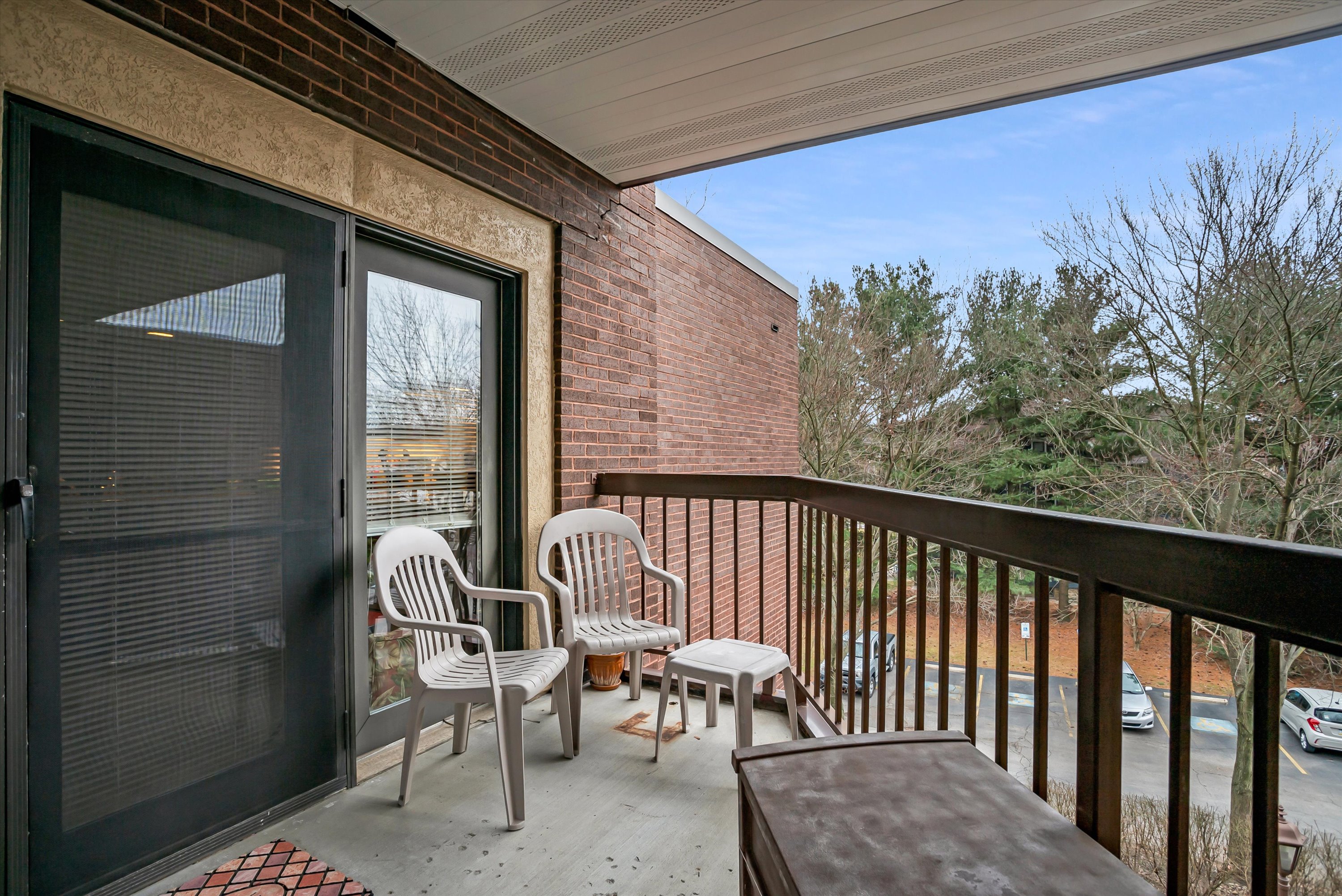 Small balcony with two white plastic chairs, a matching stool, and a railing overlooking a parking lot and bare trees.