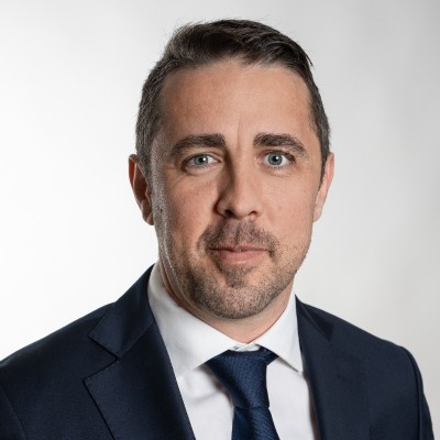 Professional headshot of a man with short brown hair and blue eyes wearing a dark suit, white shirt, and blue tie.