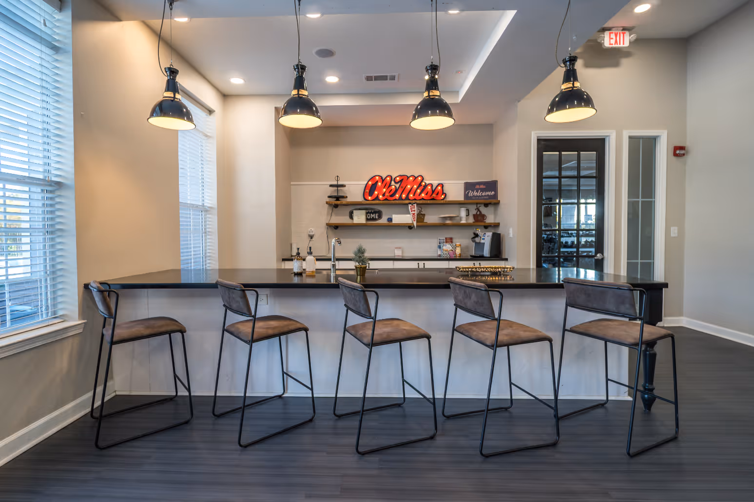 Community kitchen and bar area with bar stools, industrial pendant lighting, and a green accent wall with decorative shelving at Rev Oxford in Oxford, MS.