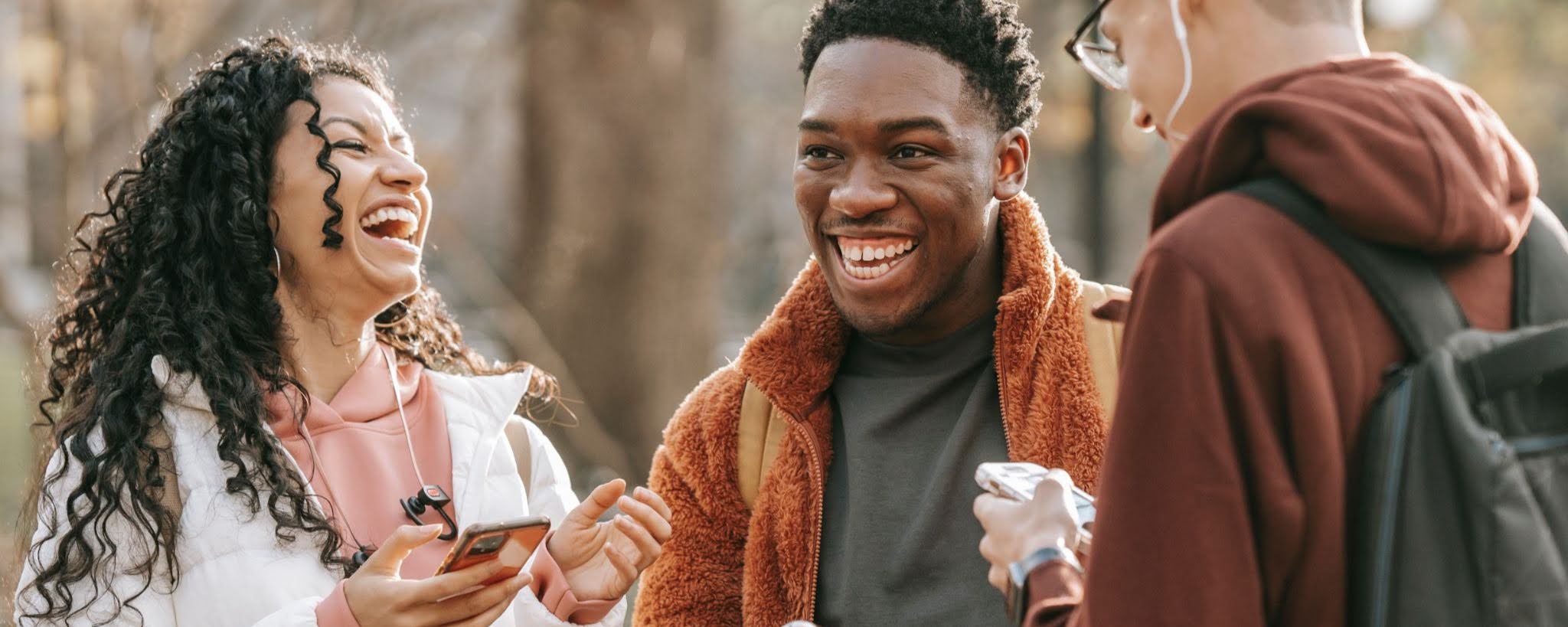 Three people laughing together outdoors on a tree-lined path, one holding a phone